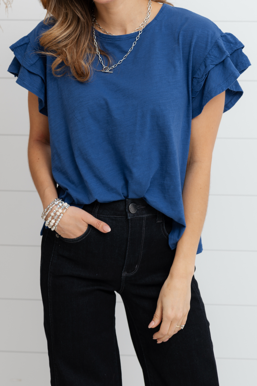 Woman wearing a blue top with ruffled sleeves against a white brick wall.