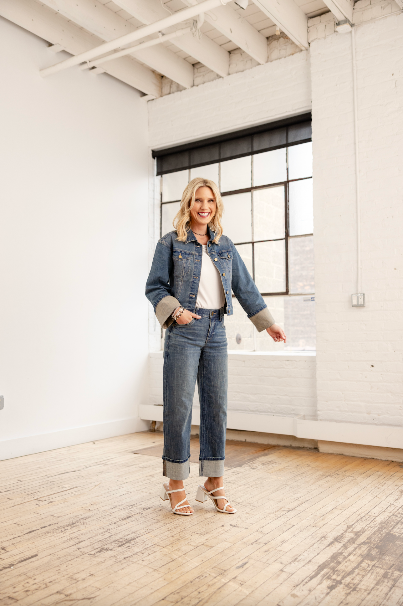 Woman wearing a denim jacket and jeans standing in a modern, minimalistic room with white walls and wooden floors.