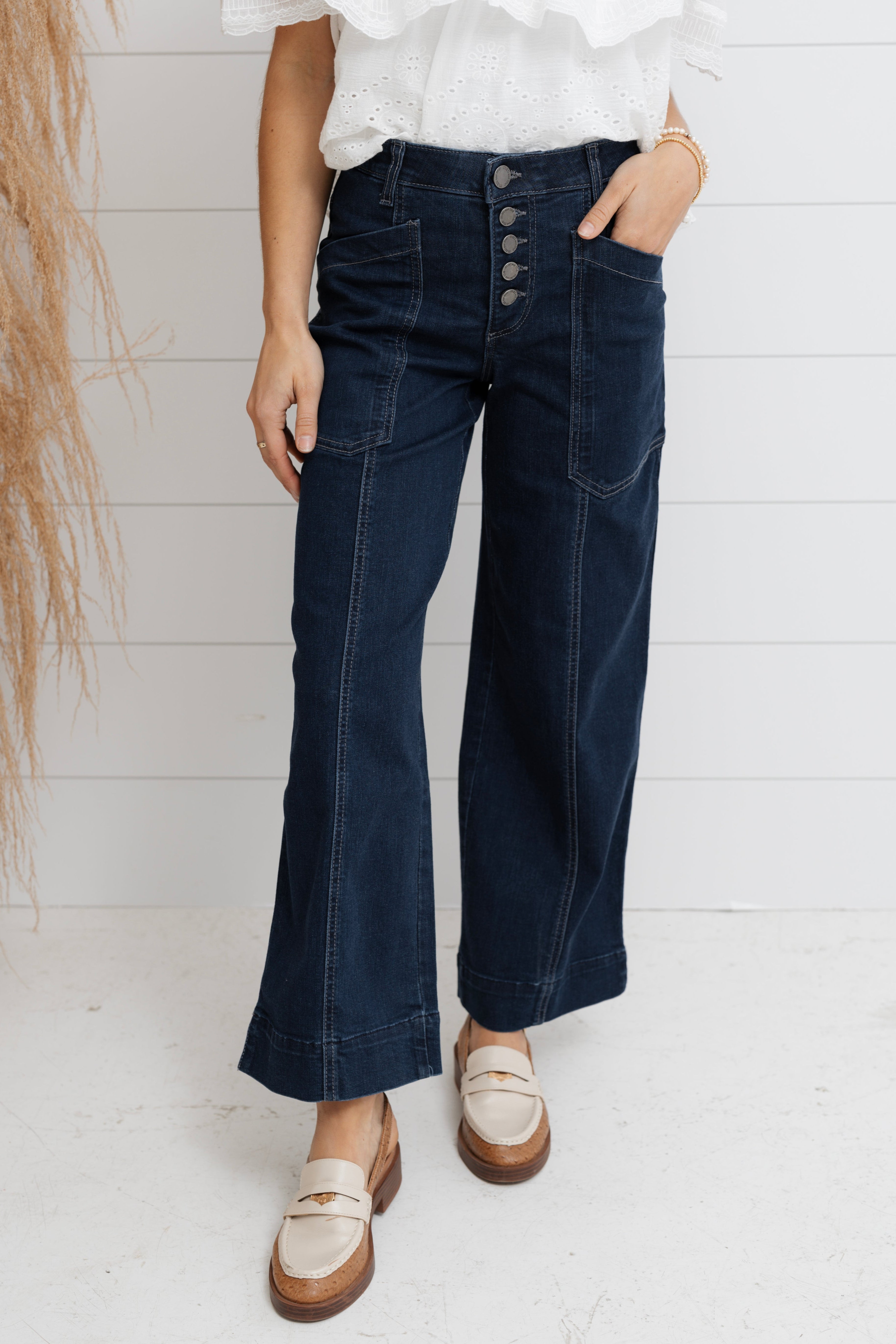 Person wearing dark blue flared jeans and a white shirt against a white wooden panel background.